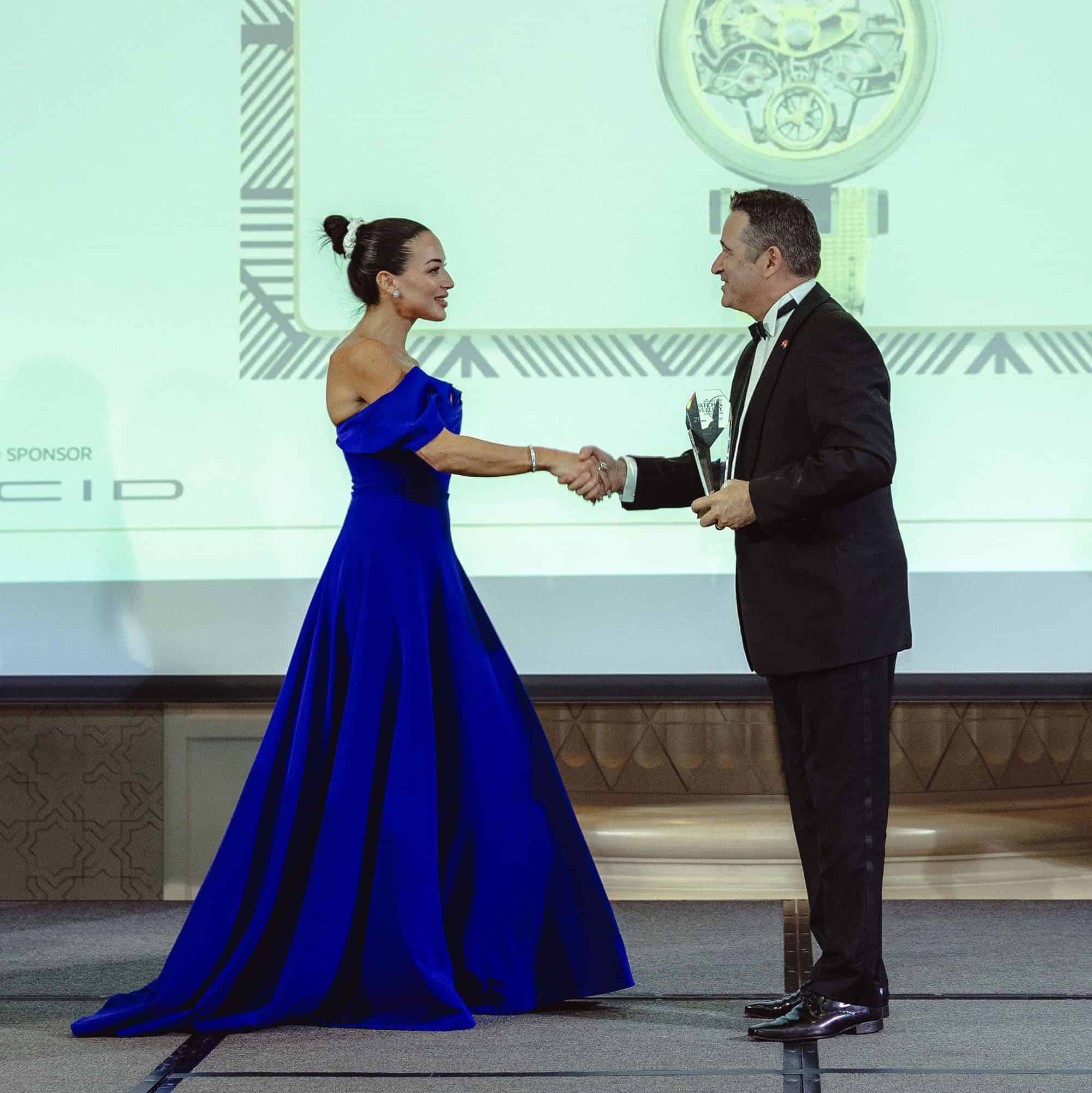 Elegant woman in a royal blue gown shaking hands with a man in a tuxedo during an award ceremony, sh.